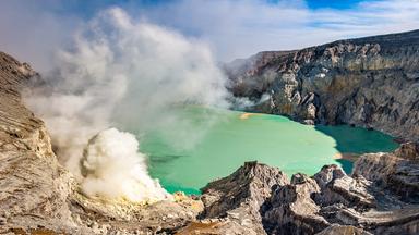 Blick auf den Krater eines aktiven Vulkans auf Java in Indonesien. Aus einer Öffnung im Vulkankrater steigt Dampf auf, daneben liegt ein türkisfarbener Kratersee in einer felsigen Landschaft.