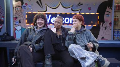 Drei junge Frauen vor einer Leuchtschrift, auf der „Welcome“ steht.