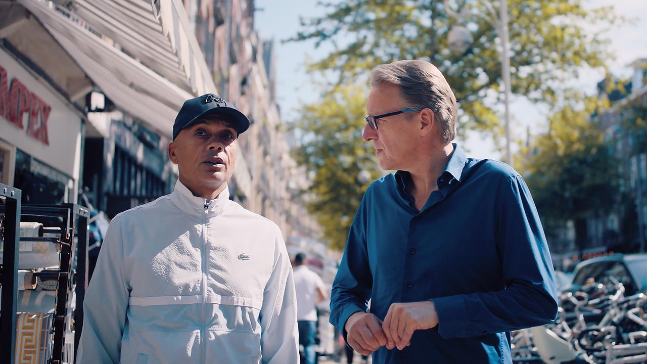 Zwei Männer zusammen auf einer Straße, der eine mit Basecap, der andere mit Brille, einander zugewandt.
