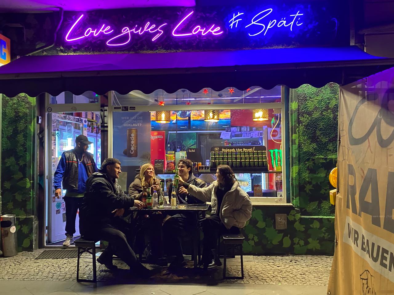 Gruppe junger Menschen sitzt vor einem mit Neonschrift beleuchteten Späti, auf der steht: „Love gives Love # Späti“.
