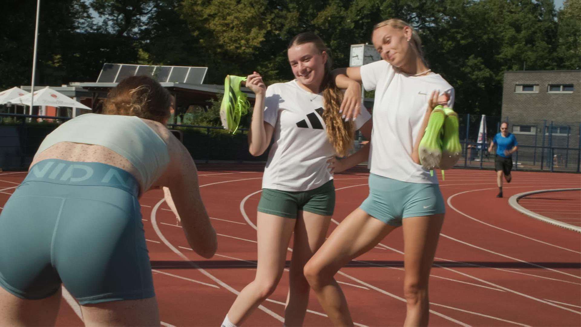 Zwei junge Sportlerinnen stehen auf einer Tartanbahn und posieren für ein Foto. Eine dritte Sportlerin fotografiert die beiden.