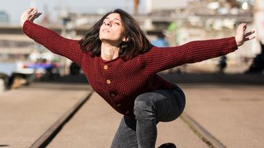 Die Tänzerin hat Oberkörper und Arme nach hinten gestreckt und schaut nach oben. Am vorderen Bildrand Ton- und Kameramann, im Hintergrund verschwommen ein Fluss mit Schiffen und Industrieanlagen. Sie trägt Straßenkleidung
