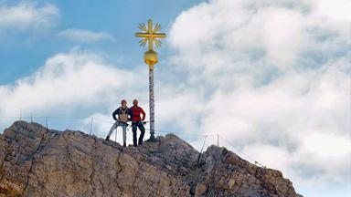 Zwei Männer stehen in Klettermontur an einem Gipfelkreuz.