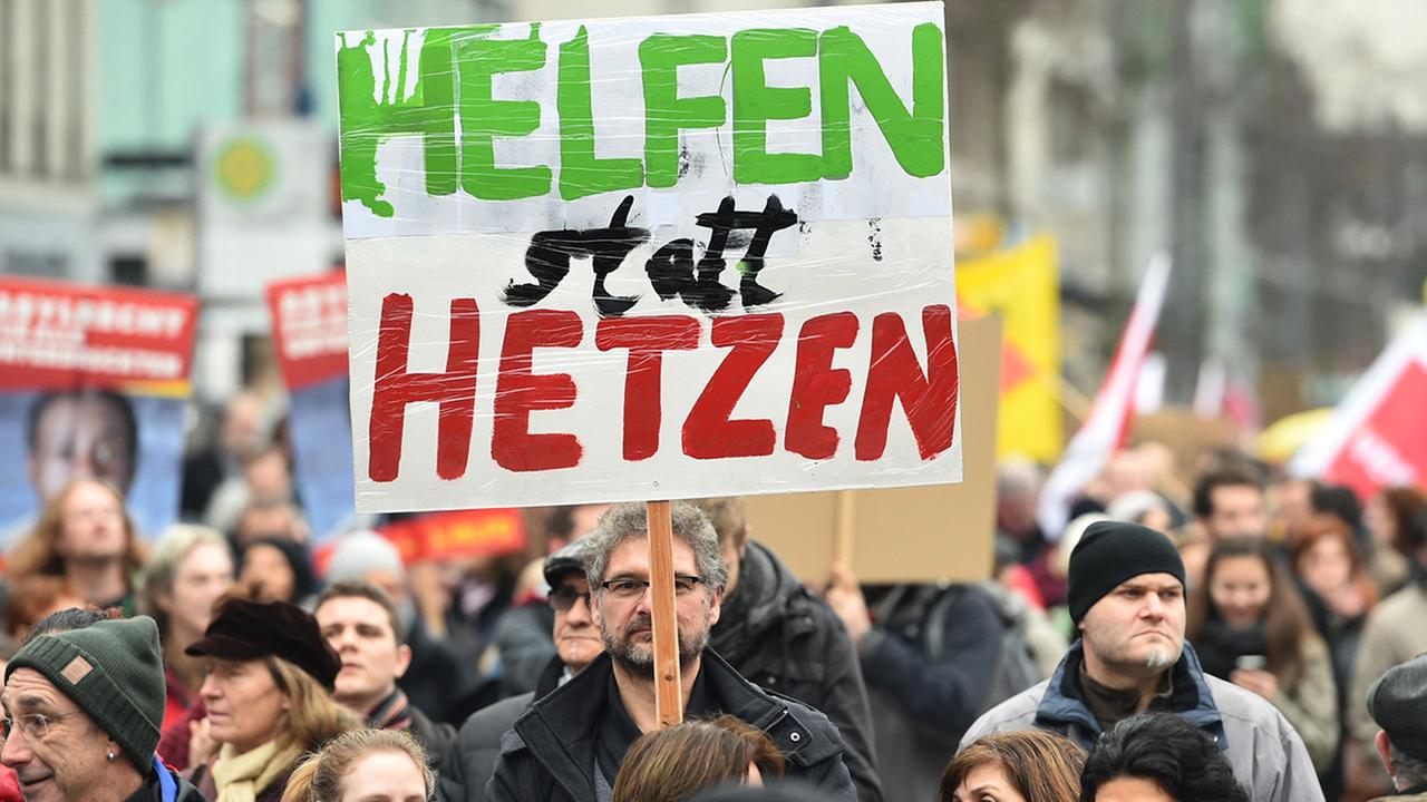 Frontalaufnahme eines Plakates mit der Aufschrift "Helfen statt Hetzen". Im Hintergund spielt sich eine Demonstration ab.