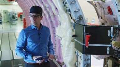 3D-Druck: Mann steht mit Ersatzteil in der Hand in einem Flugzeugrumpf, der sich im Rohbau befindet.