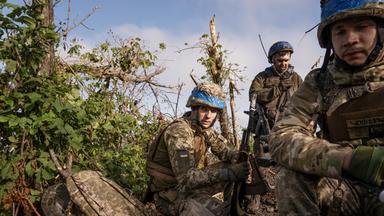 Soldaten in Camouflage kauern zwischen Büschen und Gestrüpp.