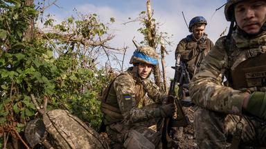 Soldaten in Camouflage kauern zwischen Büschen und Gestrüpp.