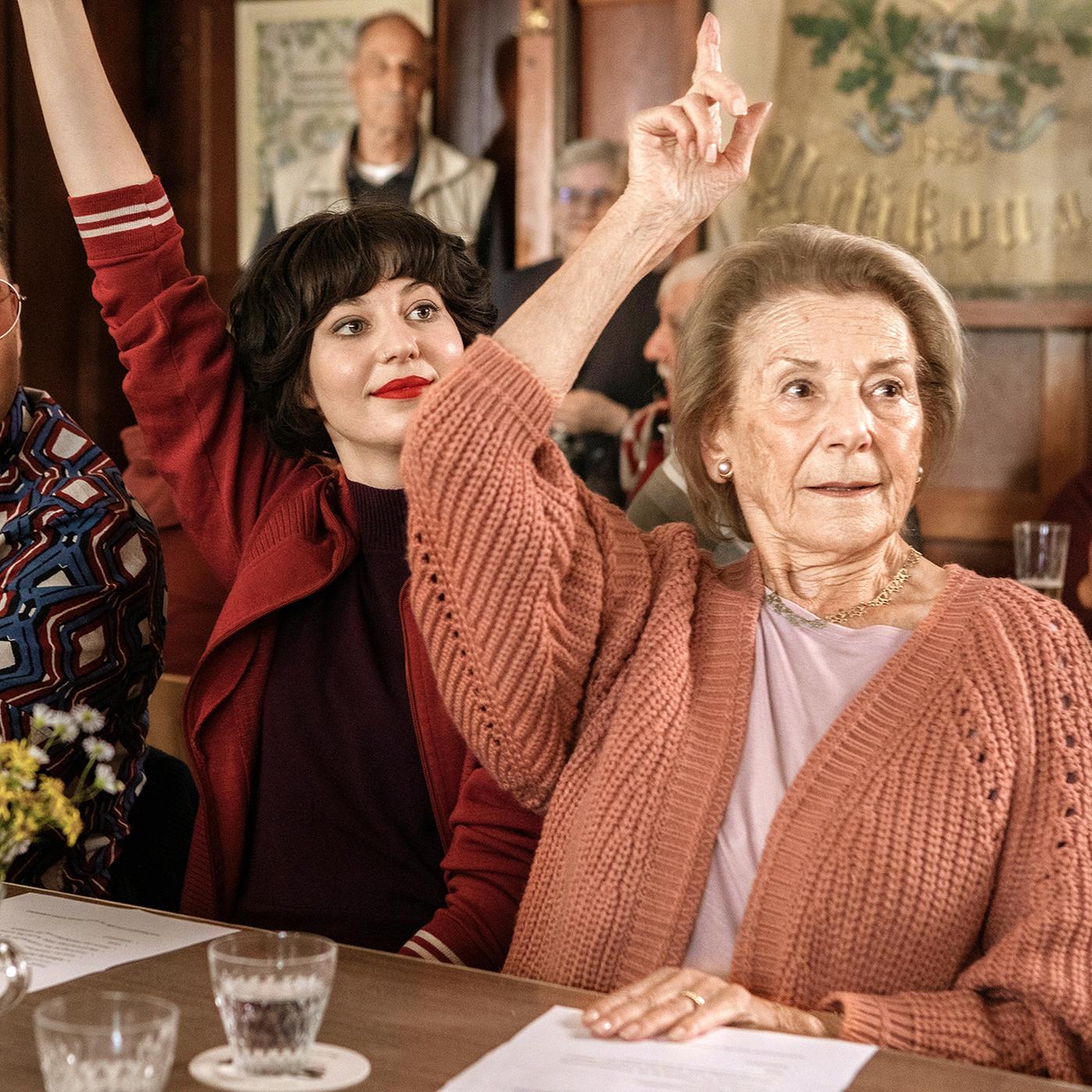 Zwei jüngere Erwachsene und eine ältere Dame heben die Hände zur Abstimmung