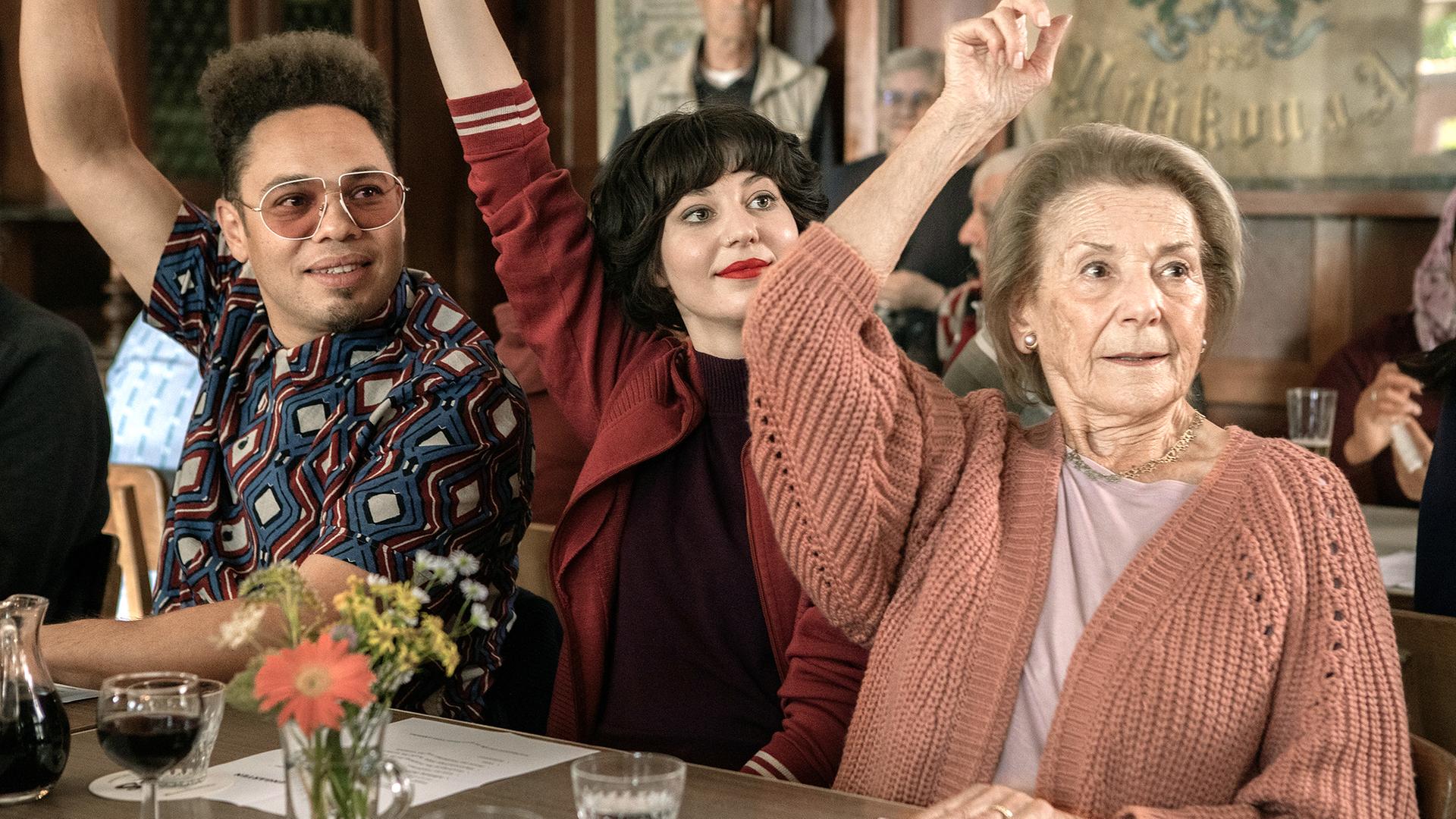 Zwei jüngere Erwachsene und eine ältere Dame heben die Hände zur Abstimmung