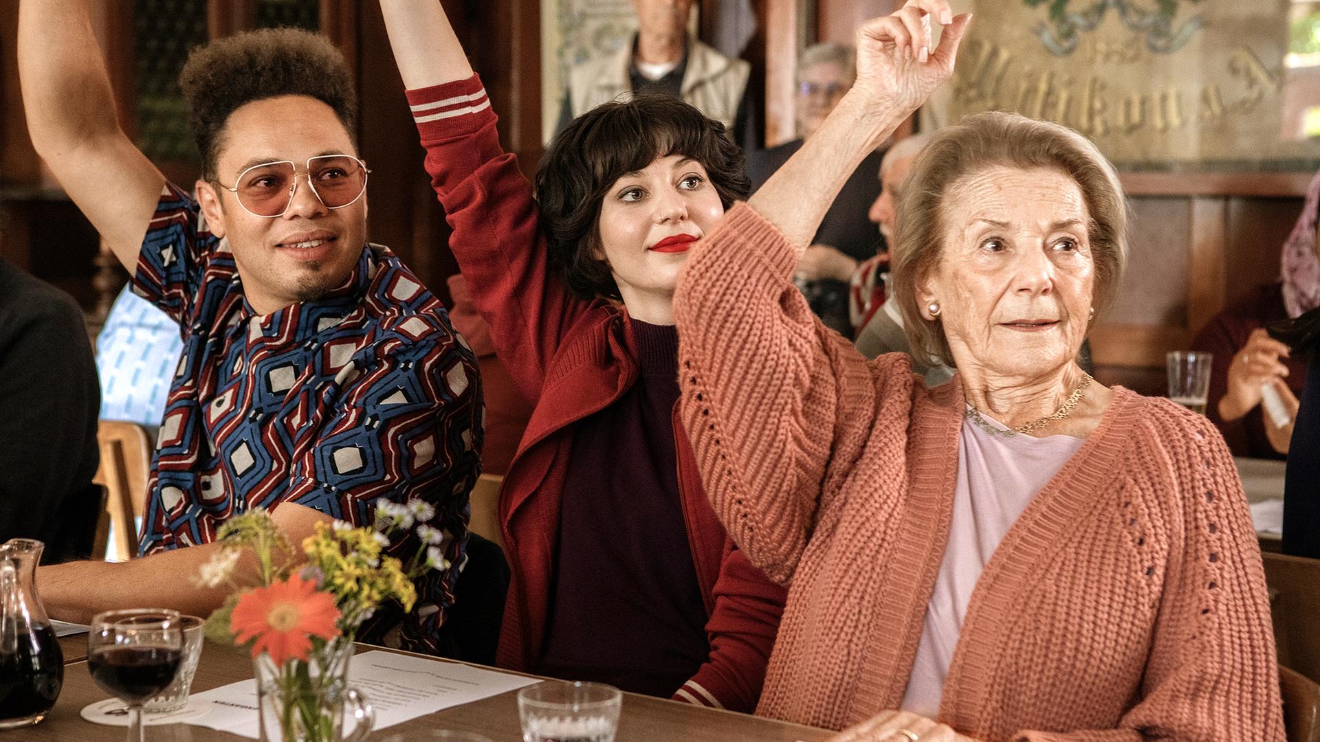 Zwei jüngere Erwachsene und eine ältere Dame heben die Hände zur Abstimmung