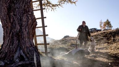 Jäger steht mit seinem Hund vor einem Hochsitz. Vor ihm ein aufgespießter toter Mann.