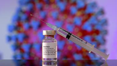 Im Vordergrund steht ein Glasfläschchen mit durchsichtigem Impfstoff, darauf lehnt eine Spritze. Im Hintergrund wabert ein Modell des Coronavirus.