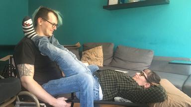 Ein Mann sitzt auf einem Stuhl. Seine Partnerin liegt auf einer Bank zwischen seinen Beinen. Ihre Füße liegen auf seinen Schultern.