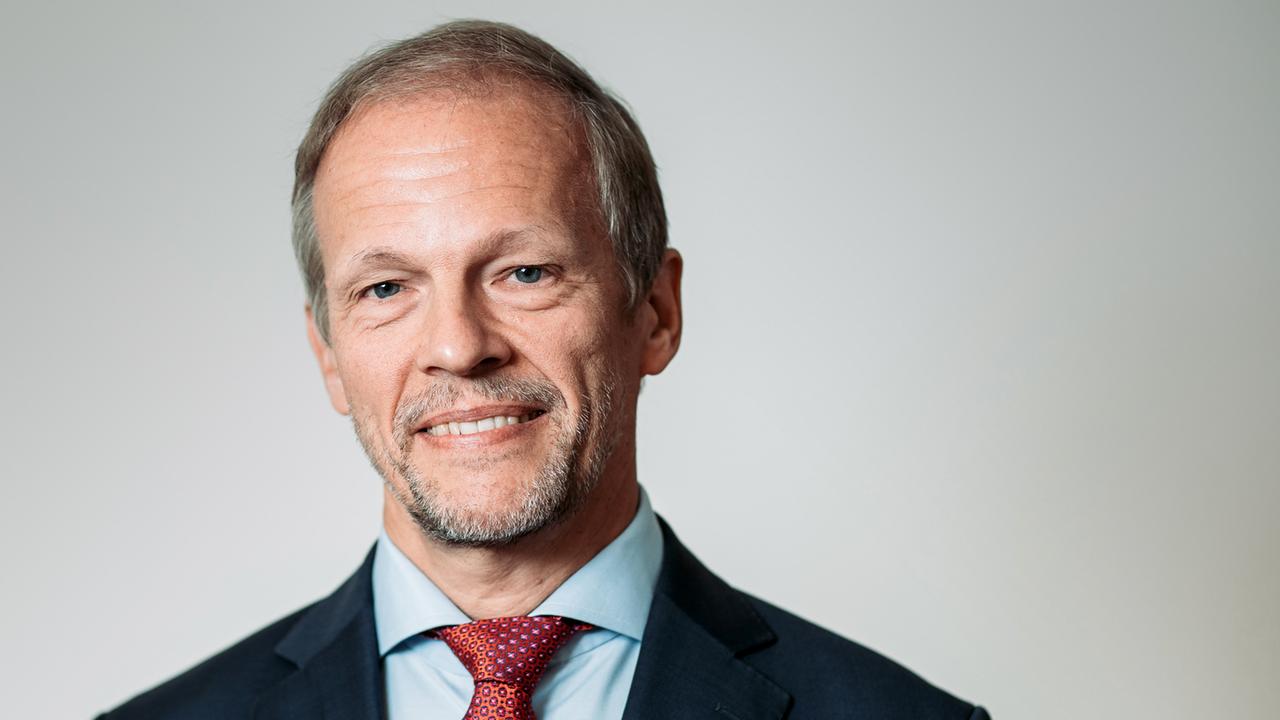 Jörg Overmann trägt einen schwarzen Anzug mit roter Krawatte und lächelt mit leicht grauem Bart in die Kamera