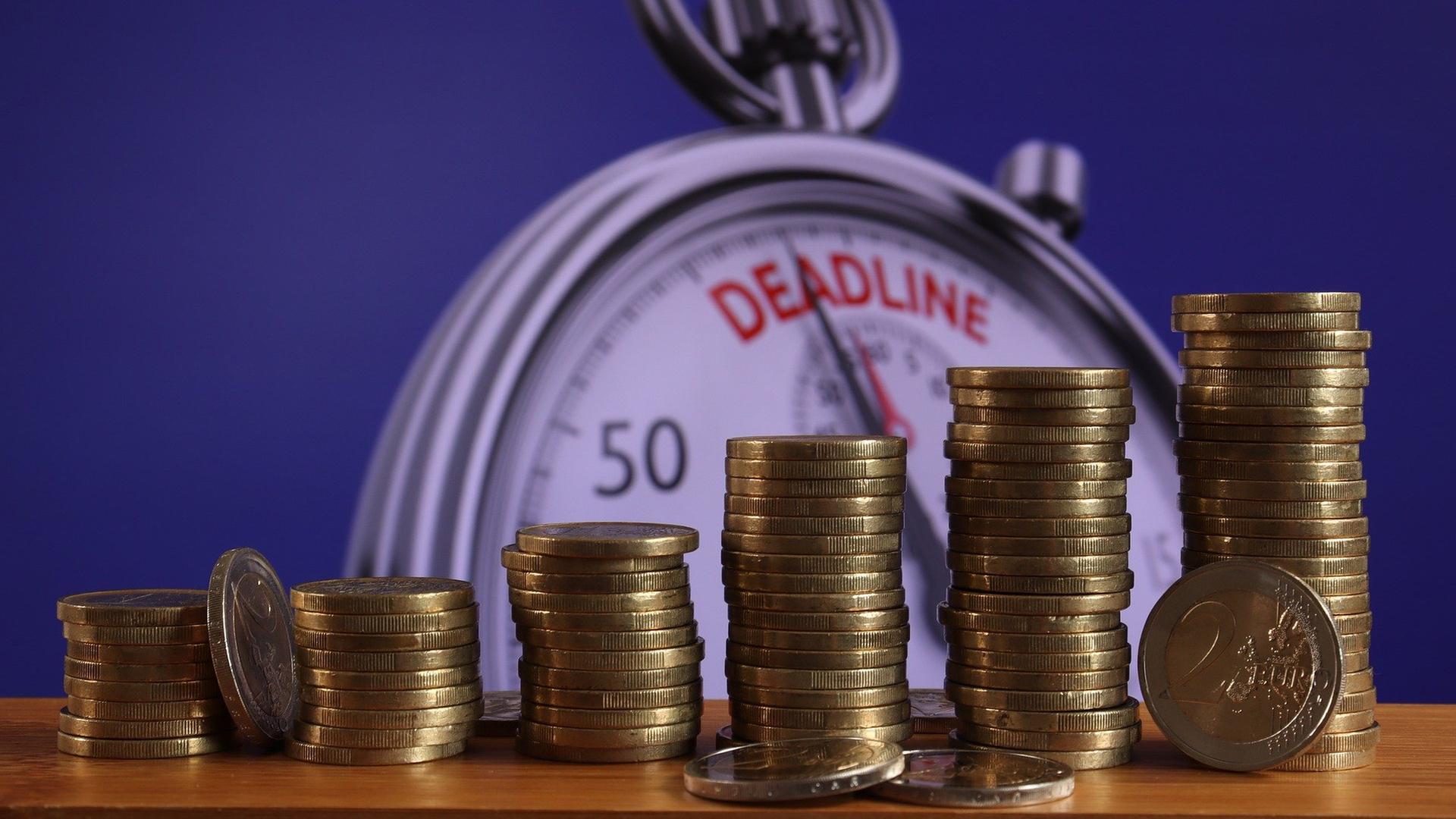 Münzen aufeinandergestapelt vor einer Uhr