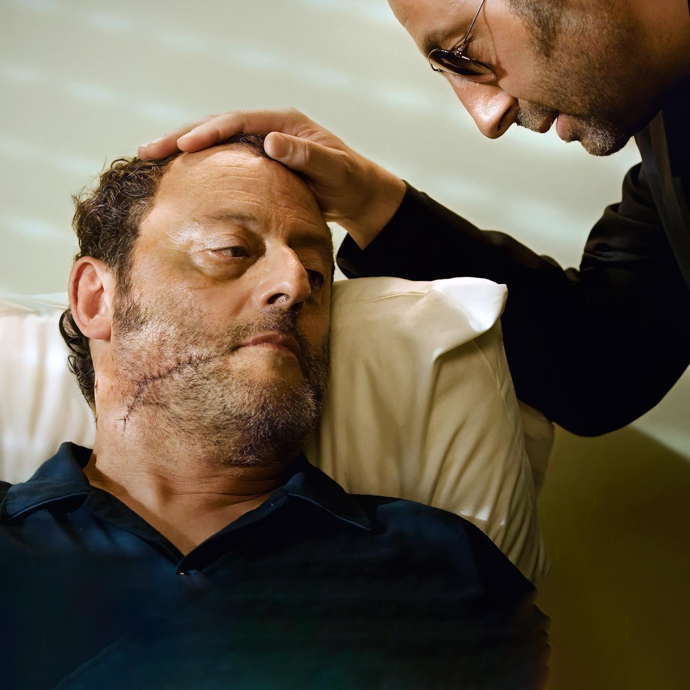 Ein Mann beugt sich über eine anderen Mann mit einer großen Narbe im Gesicht und legt seine Hand auf seinen Kopf.