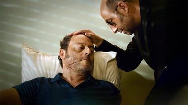 Ein Mann beugt sich über eine anderen Mann mit einer großen Narbe im Gesicht und legt seine Hand auf seinen Kopf.