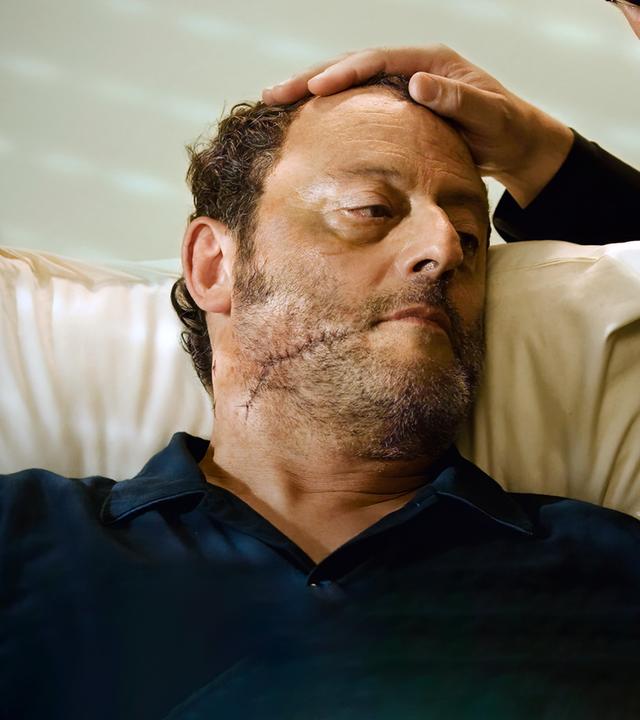 Ein Mann beugt sich über eine anderen Mann mit einer großen Narbe im Gesicht und legt seine Hand auf seinen Kopf.