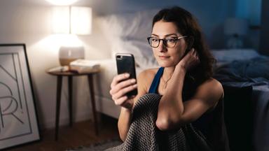 "makro: Liebe auf den ersten Klick - Milliardenmarkt Onlinedating": Eine Frau sitzt mit angezogenen Knien an die Seite ihres Bettes angelehnt auf dem Boden. und schaut in ihr Smartphone.