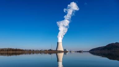 Wasserdampf steigt aus dem Kühlturm des Atomkraftwerks Isar 2. Im Hintergrund strahlend blauer Himmel, im Vordergrund spiegelt sich die gesamte Szenerie auf der glatten Oberfläche der Isar.