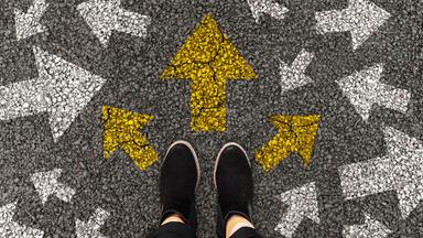 Füße auf einer Straße auf der Pfeile in verschiedene RIchtungen aufgemalt sind.