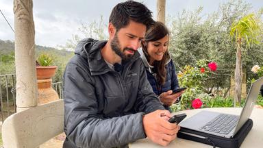 "makro: New Work - Die Zukunft der Arbeit": Jan Navel, ein großer dunkelhaariger Mann mit Bart und Greta Navel, eine Frau mit langen dunklen Haaren, sitzen mit einem Notebook in einem Pavillon und schauen suchend auf ihre Handys. Im Hintergrund ein Garten und Bäume.