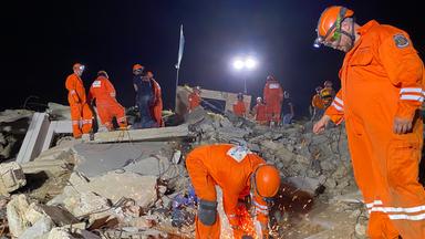Männer in orangefarbenen Schutzanzügen suchen unter Trümmern nach Überlebenden.