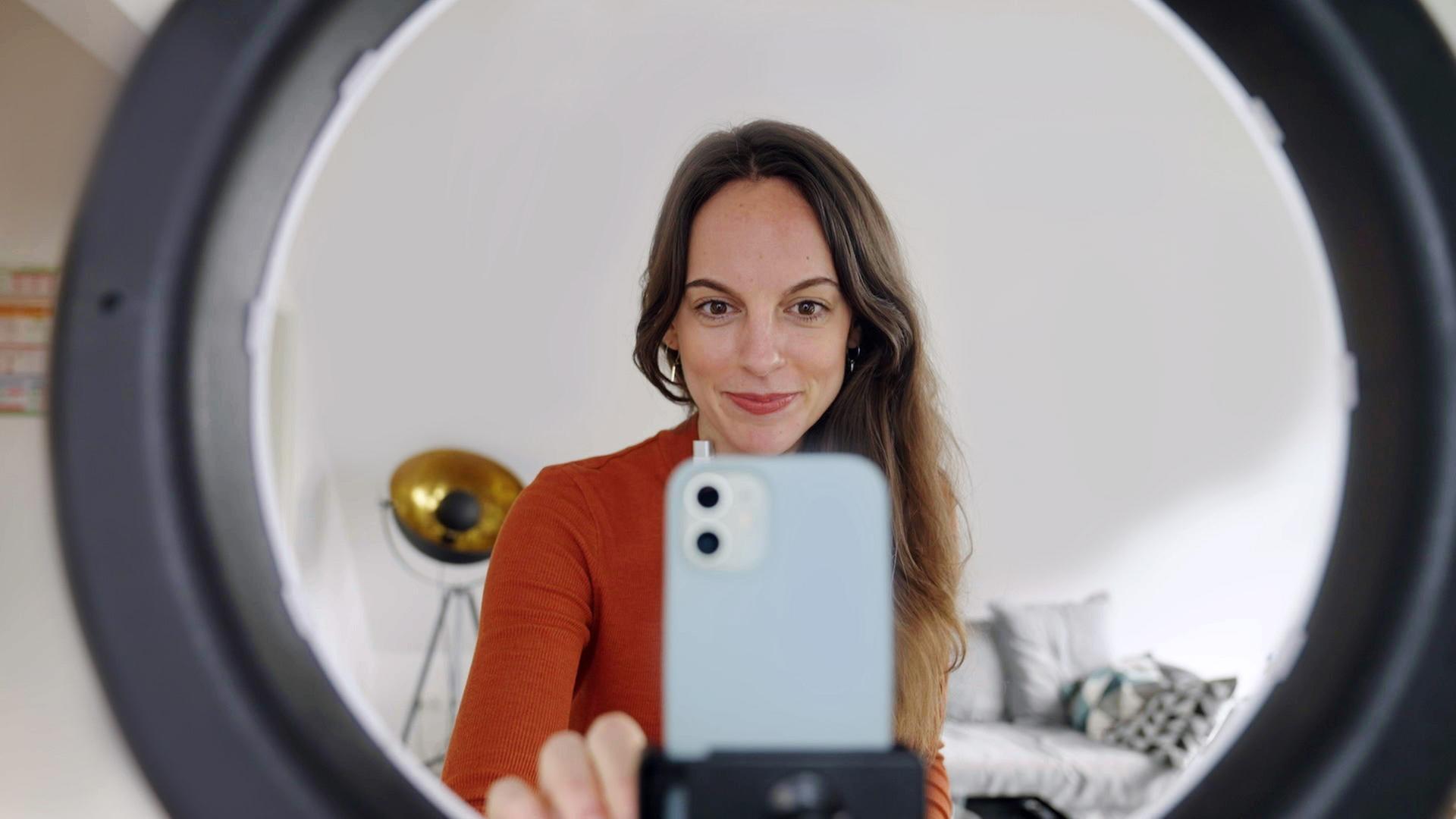 "makro: This is TikTok - Stars und Hype in Hochkant": Gianna Bacio sieht man durch ein Ringlicht. Mit ihrer rechten Hand stellt sie die Position eines Handys ein, das sich in der Mitte des Ringlichts befindet.