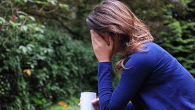 Frau sitzt mit Tasse draußen und hält sich mit einer Hand den kopf. Sie scheint deprimiert zu sein oder Schmerzen zu haben.