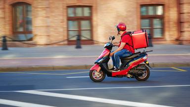 Ein Lieferdienstfahrer fährt mit seinem Moped und seiner roten Tragetasche auf dem Rücken durch die Straßen.