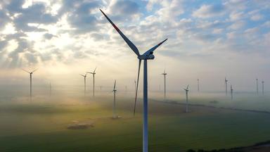 Das Luftbild einer Drohne zeigt die Morgensonne hinter Wolken über der in Nebel gehüllten Landschaft an einem Windenergiepark in Brandenburg.