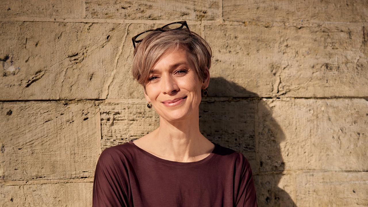 Tatjana Schneider, Architektin, steht vor einer Mauer. Sie hat kurze, hell blonde Haare und trägt eine Brille.