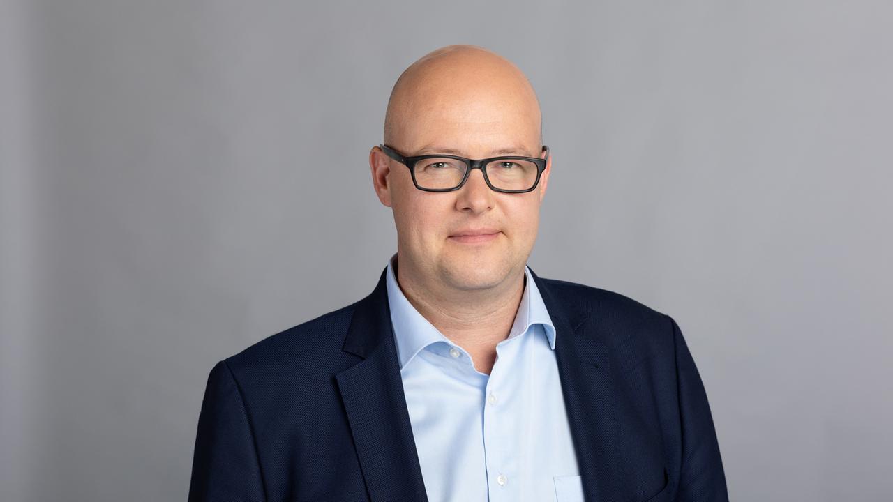 Leonhard Dobusch, Wirtschaftswissenschaftler, steht vor einer grauen Wand. Er trägt ein dunkles Jacket, eine dunkle Brille und hellblaues Hemd.