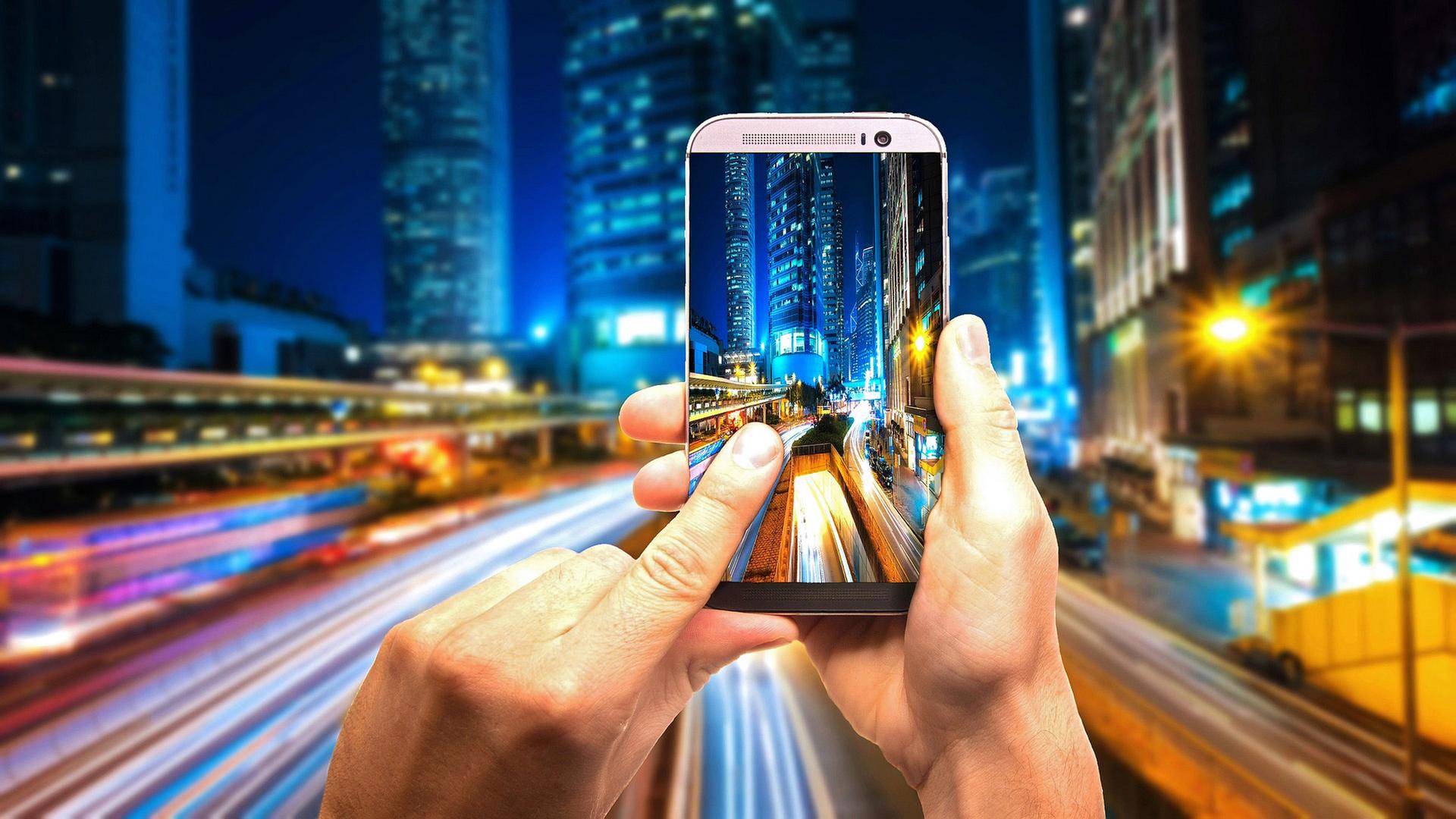 Nächtlicher Straßenzug, im Vordergrund eine Hand mit Handy