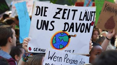 Viele Demonstrierende halten Plakate für mehr Klimaschutz in die Luft. Im Mittelpunkt erkennt man ein großes Schild, mit der Aufschrift "Die Zeit läuft uns davon". Außerdem ist eine explodierende und brennen Weltkugel auf das Plakat gemalt.