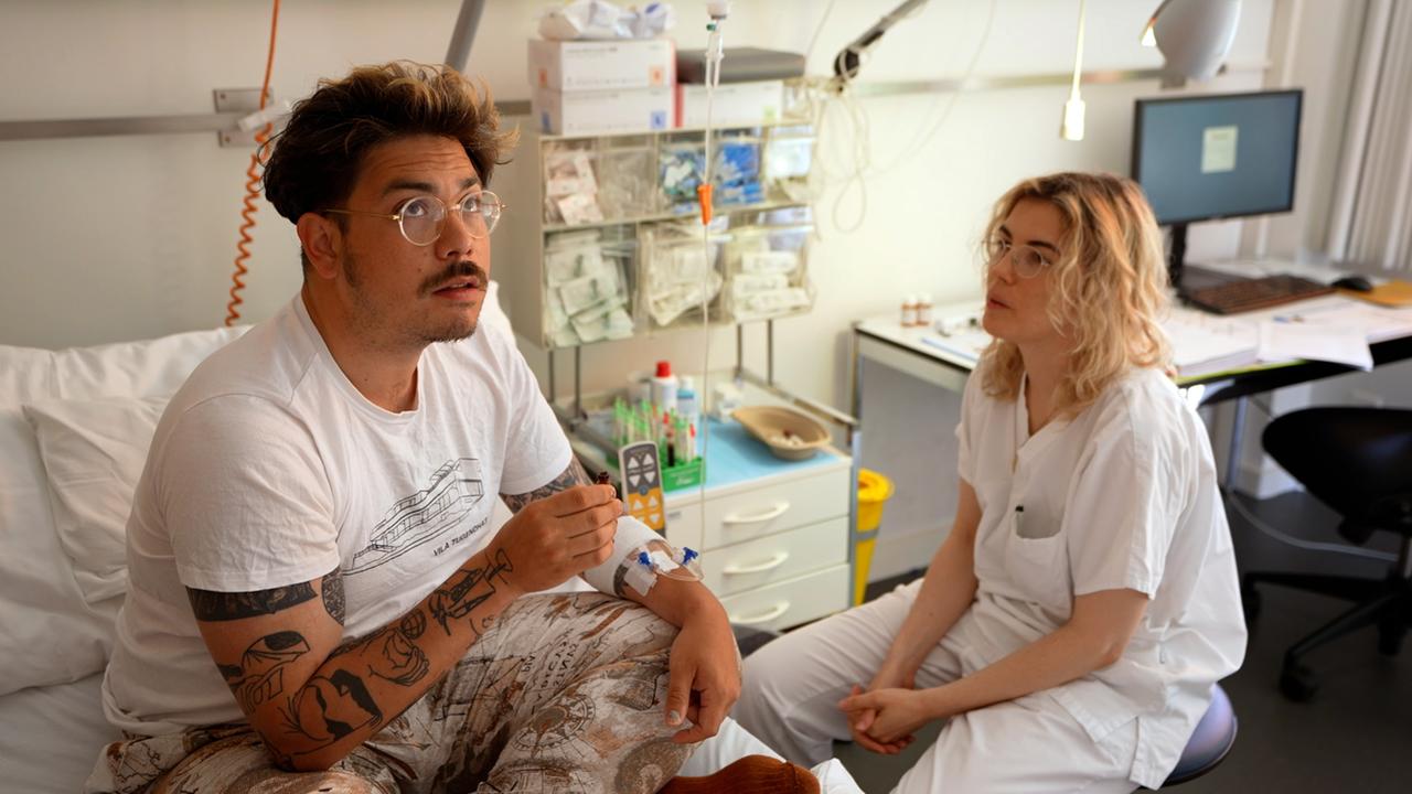 Mann in Krankenhausbett mit Kanüle am linken Arm nimmt unter medizinischer Betreuung einer neben dem Bett sitzenden Frau Medizin ein.