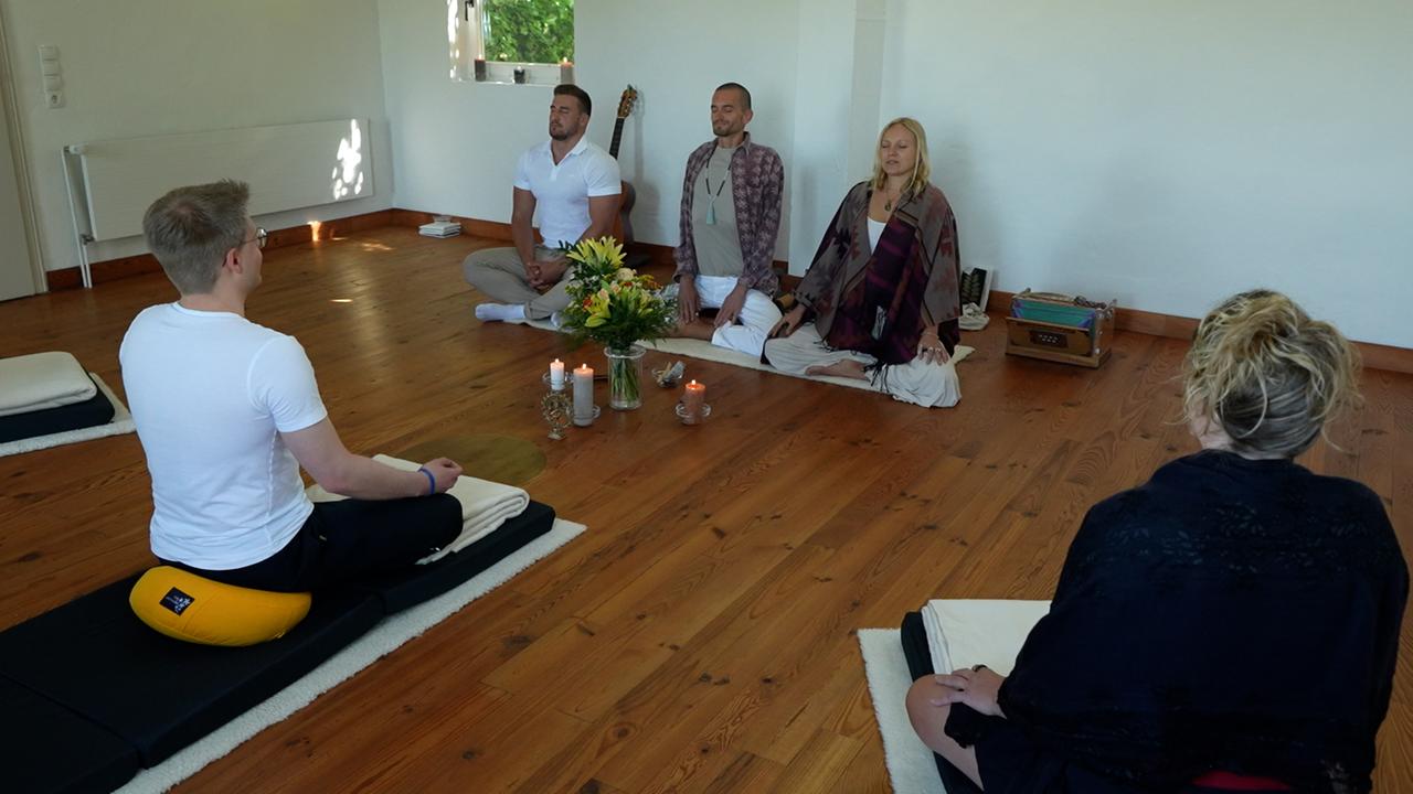Fünf Personen auf Yogamatten sitzen im Schneidersitz in einem mit Holzboden ausgelegten Raum. Zwischen ihnen drei brennende Kerzen und ein Blumenstrauß