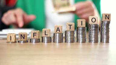 Auf einem Tisch sind Münzen auf verscheiden hohen Münzstapeln aufgestapelt, darauf sind Holzwürfel mit der Inschrift Inflation. Im Hintergrund tippt eine Person auf einem Taschenrechner und hält Geldscheine in der Hand.