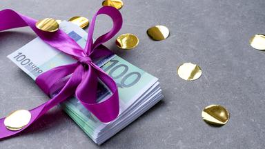 Ein Stapel 100-Euro-Scheine mit lila Schleife liegen auf einem grauen Untergrund.