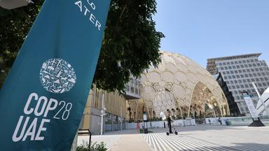 Logo COP28 auf einem Plakat, daneben ein Blick auf ein Gebäude