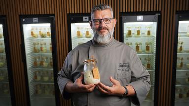 Ein Mann in grauer Kochjacke hält ein Einmachglas mit Sauerteig in der Hand.