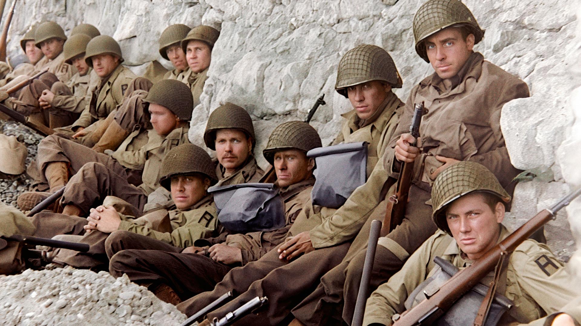 Ein gutes Dutzend amerikanischer Soldaten mit Gewehren und Helmen kauern vor einem weißen Felsen.