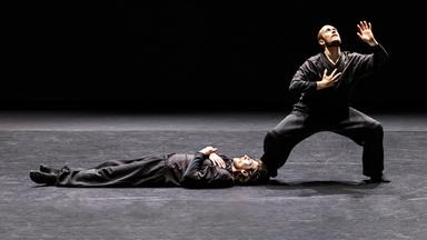 Zwei Tänzer in schwarzer Kleidung auf einer Bühne. Einer liegt flach auf dem Boden.