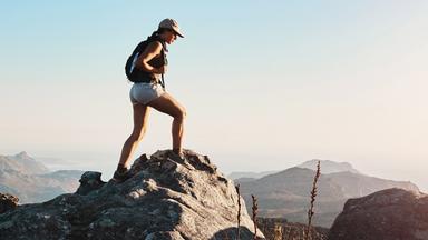 Vor blauem Himmel steht eine Frau auf einer Bergspitze und schaut ins Tal.