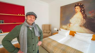 Max Schmidt steht links von einem Hotelbett. Über dem Bett hängt ein großes Gemälde der Kaiserin Sisi.