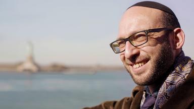 Rabbi Akiva Weingarten (39) lächelt im Vordergrund, während im Hintergrund die Freiheitsstatue und der Hudson River in New York unscharf zu sehen sind.