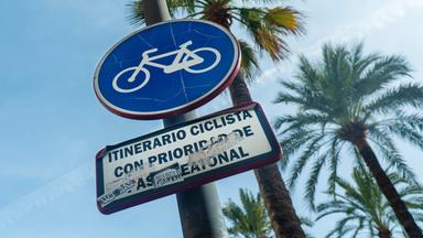 Im Vordergrund ist ein blaues, rundes Schild, auf dem ein weißes Fahrrad zu sehen ist. Dieses Schild steht für „Sonderwege für Fahrradfahrer“. Unter dem runden Schild ist noch ein rechteckiges Hinweisschild auf Spanisch „Mit Priorität für Fußgänger“ angebracht. Im Hintergrund sieht man den strahlend blauen Himmel und Palmen.
