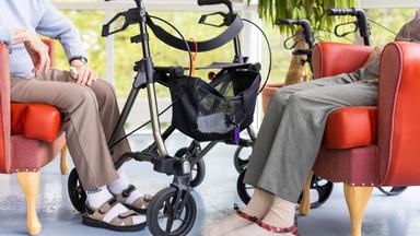 Alte Menschen sitzen auf Sesseln, dazwischen ein  Rollator