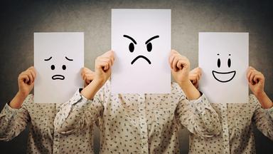 Eine Person steht vor einer Wand und hält verschiedene Smileys vors Gesicht.