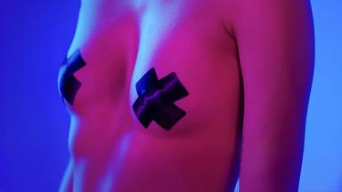 Nackter Oberkörper einer Frau. Die Nippel sind mit Klebeband in Form eines „x“ abgeklebt. Blau-pinke Farbstimmung.
